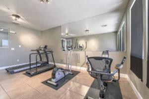 Interior Fitness center, treadmills, elliptical, and stationary bike. Tan tile flooring, gray walls, wall mirrors in front of exercise equipment.
