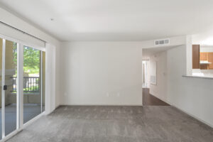 Interior unit living room, gray carpeting in living room, sliding glass door to patio area left side of photo, wood-like floored hallway leading to bedroom and bathroom right side of photo.