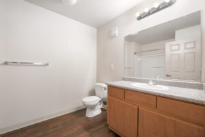 Interior unit bathroom, wood like floors, large vanity mirror, faux granite countertop, towel rack, bathtub/shower, white walls.